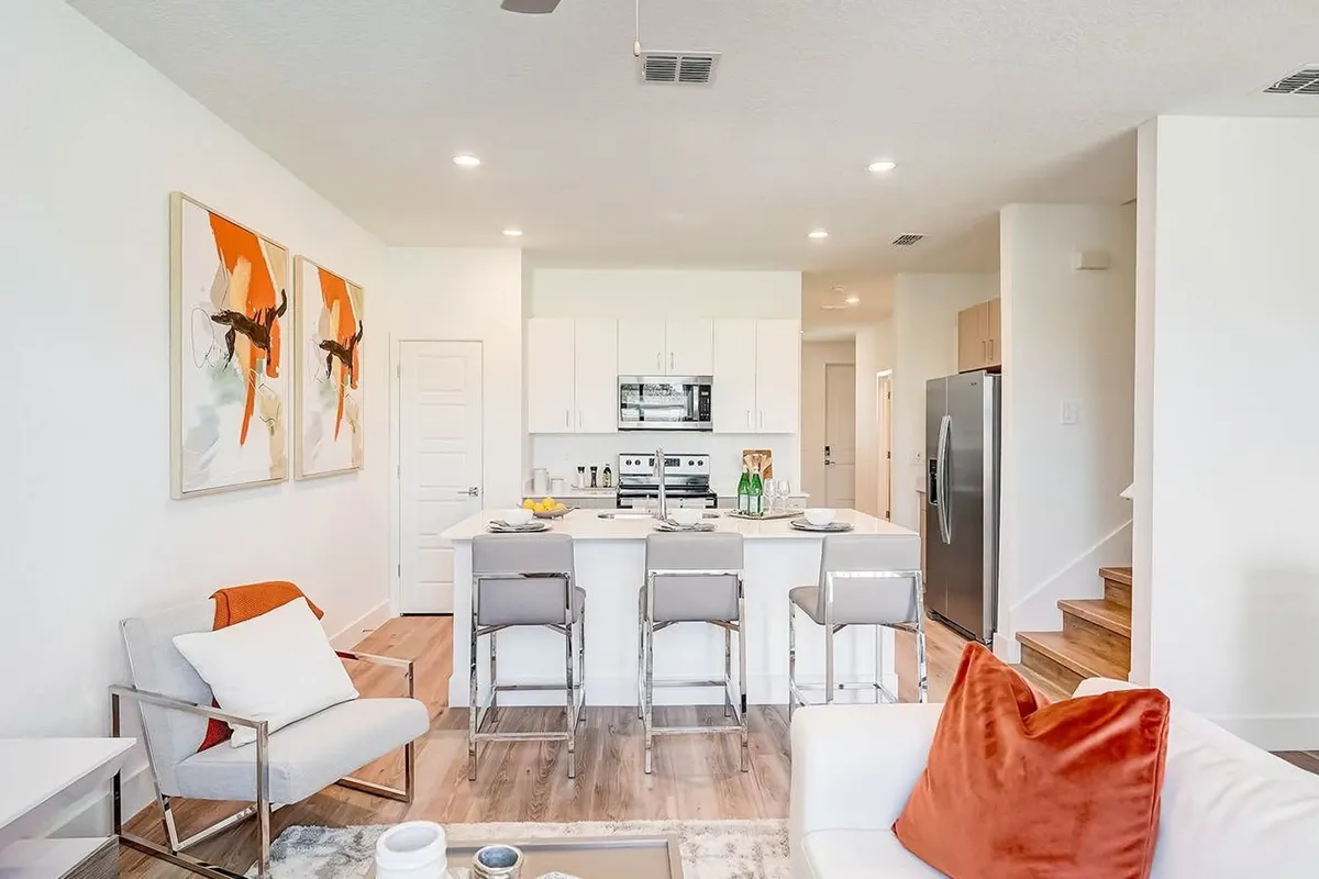 View from the living room into the kitchen, with three bar stools at the island, the range under a microwave, a stainless refrigerator and the stairs