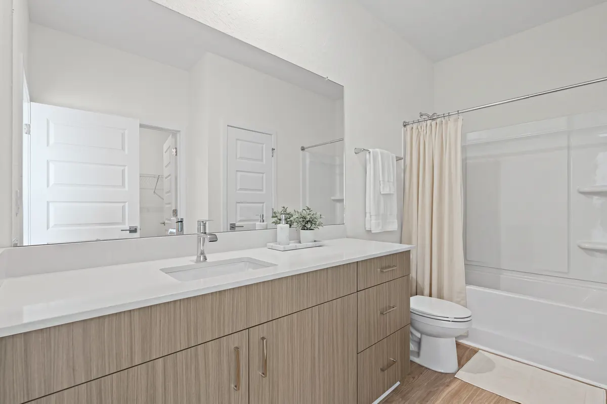 Amber bathroom with a long wood vanity and a wide mirror, white towels on the towel bar, a marble tray with a soap dispenser and two small plants, an ivory shower curtain gathered at the end of the tub and a bath mat. Virtually staged.
