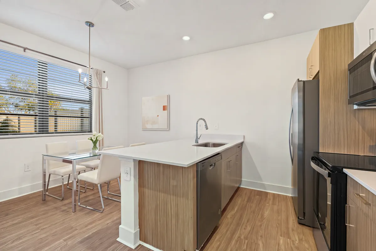 Amber kitchen with an island, stainless appliances and light cabinetry, and a glass dining table and chairs by the window. Virtually staged.