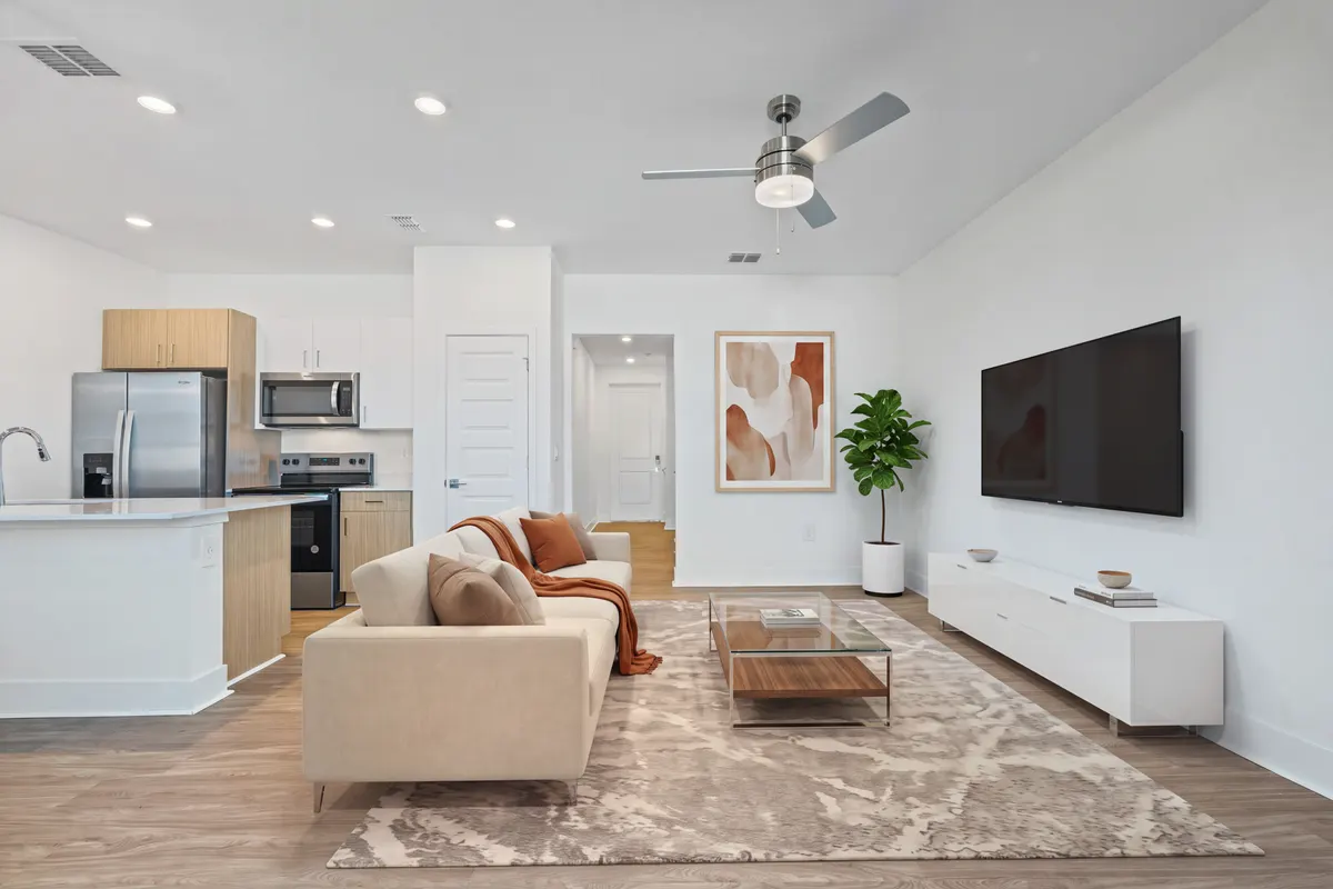 Amber living room with a wall-mounted TV over a white media console, a sofa, a glass coffee table, a rug and abstract art, looking through to the kitchen. Virtually staged.