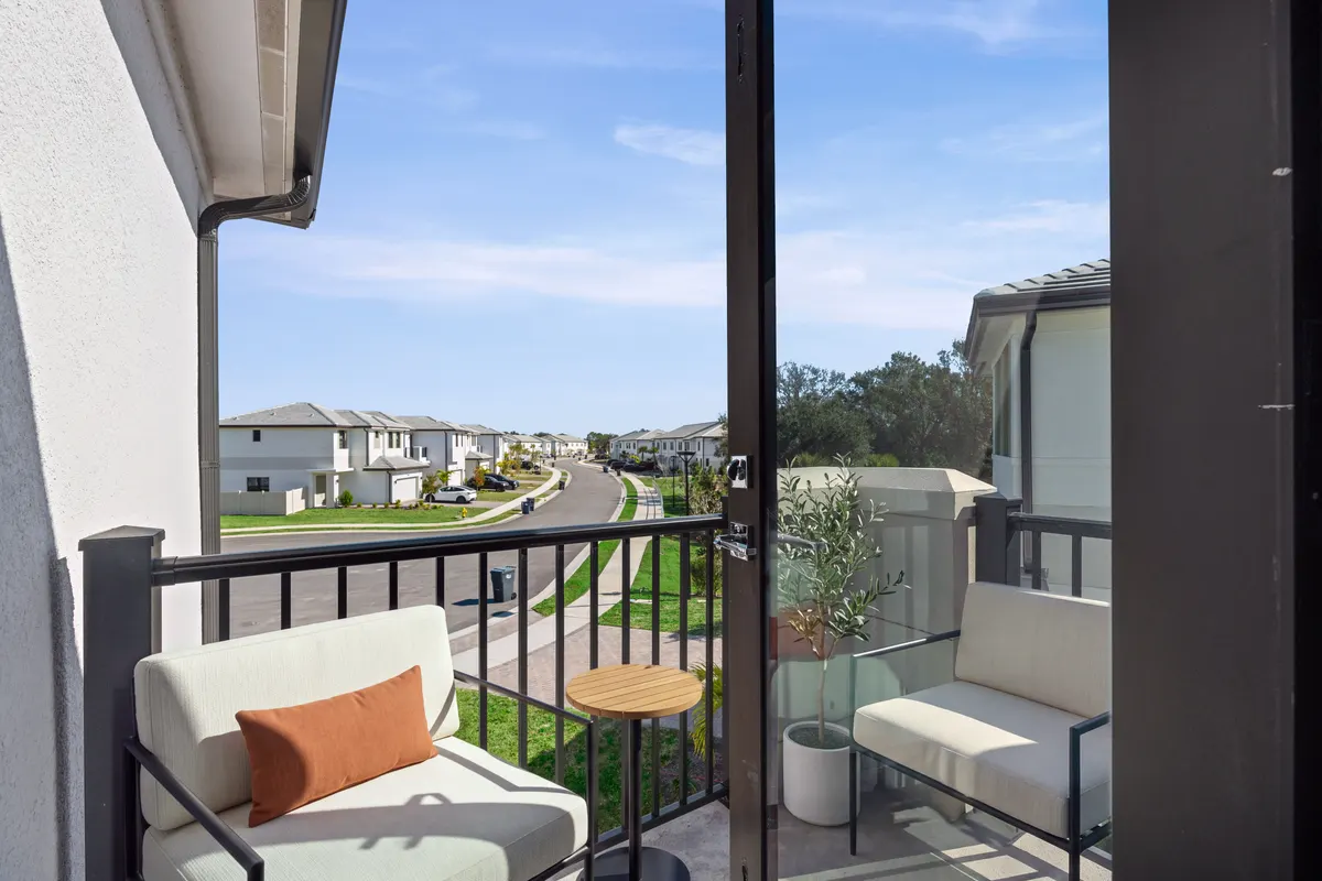 Maple second-floor balcony with two ivory-cushioned lounge chairs, a round teak side table and a potted olive tree, looking out over the street. Virtually staged.