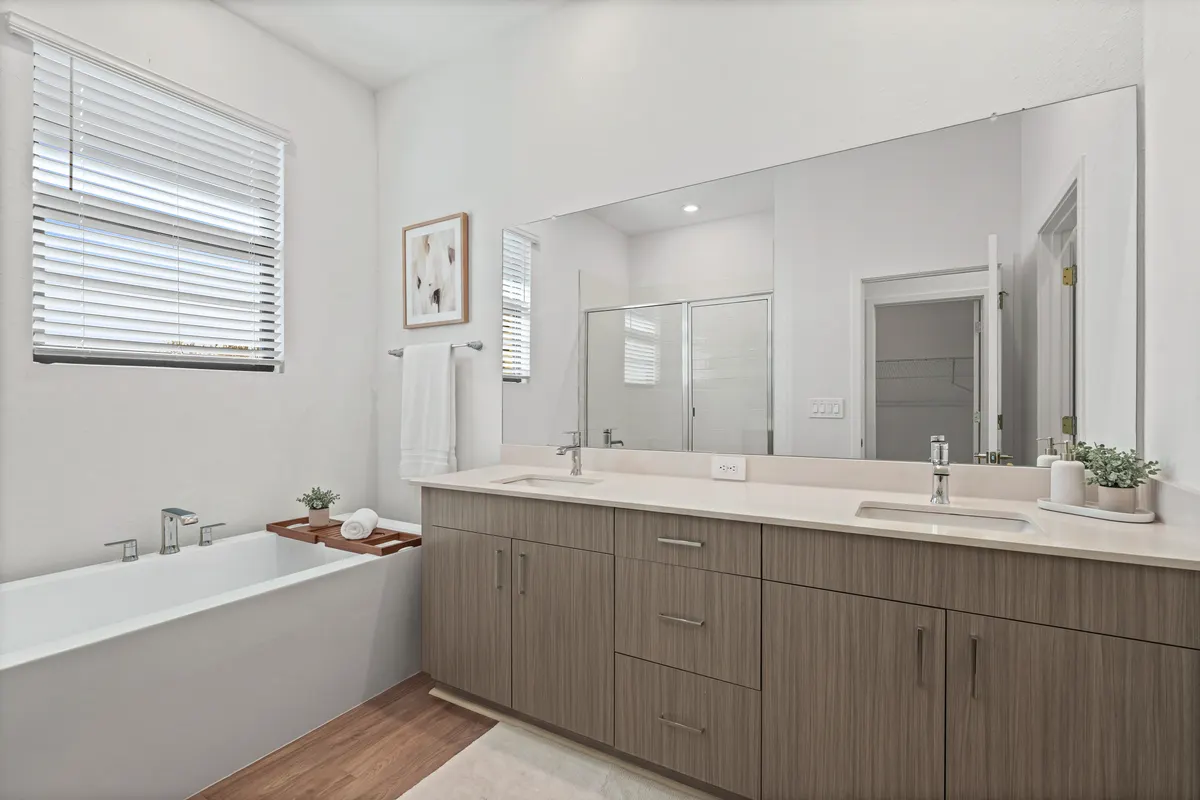 Maple primary bathroom with a wooden bath tray across the freestanding tub, a towel on the bar below a framed print, and a double vanity with a soap tray and a bath mat. Virtually staged.