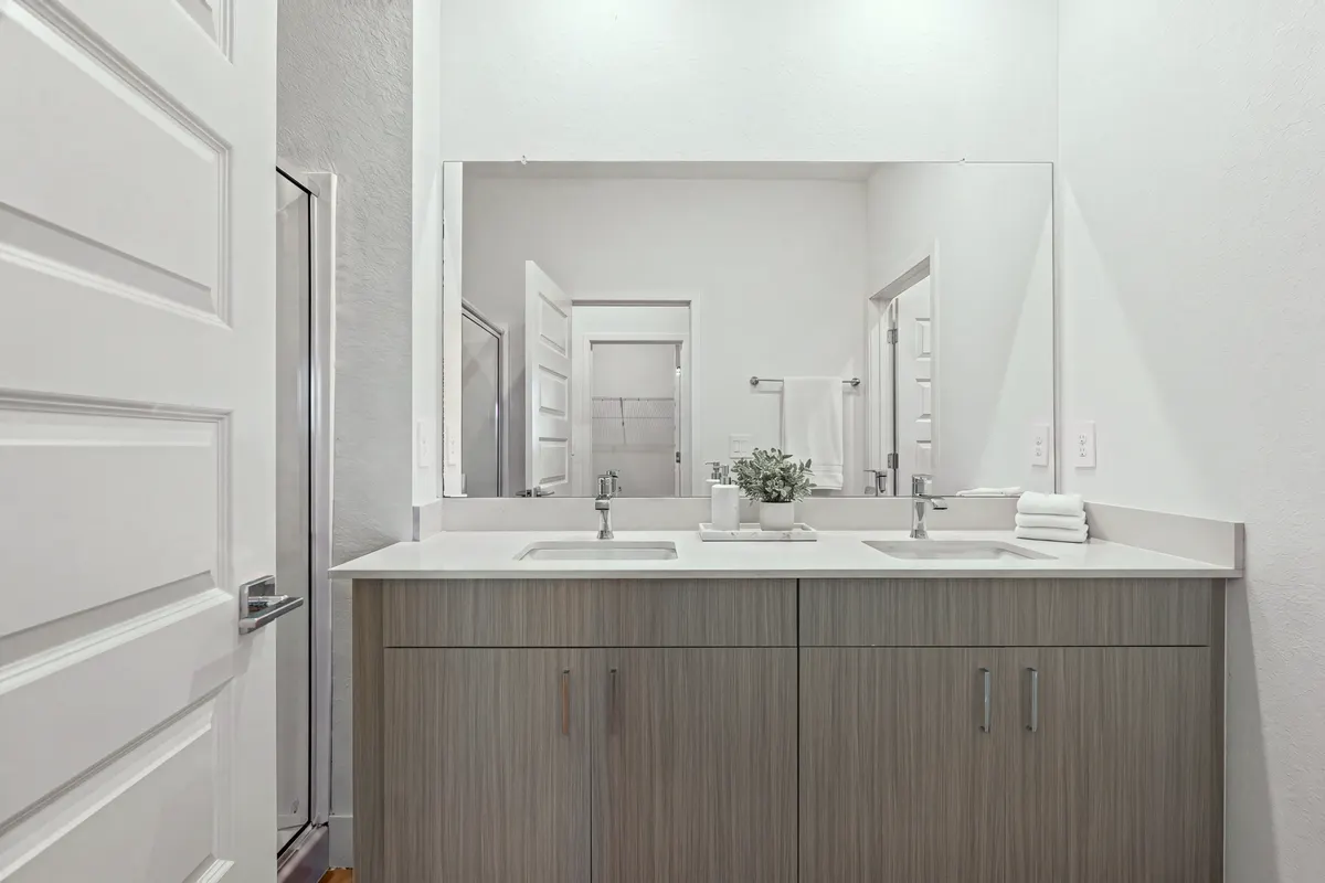 Opal bathroom with a double vanity and a wide mirror, a marble tray with a soap dispenser and a small plant between the sinks and folded white hand towels. Virtually staged.