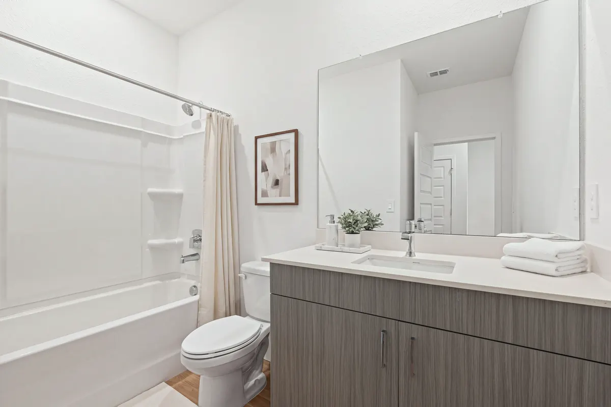 Opal hall bathroom with an ivory shower curtain gathered at the end of the tub, a small abstract print above the toilet, a vanity with a marble tray, soap dispenser and small plant, folded white towels and a bath mat. Virtually staged.